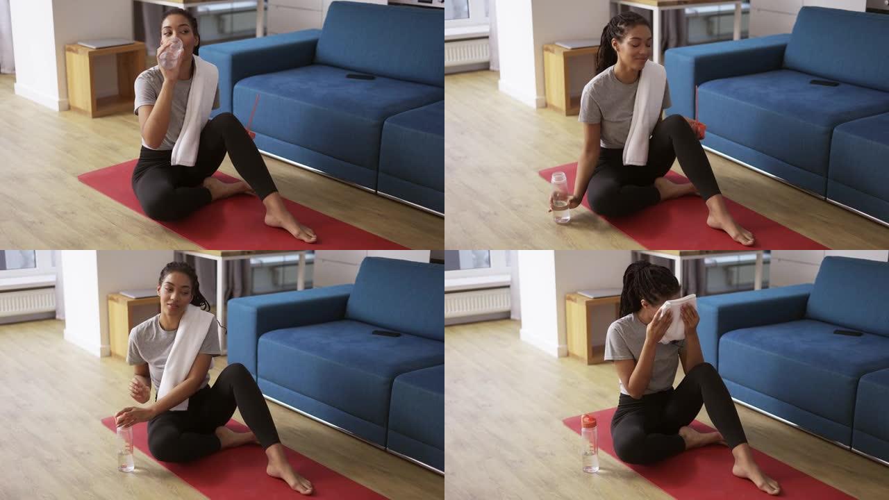 运动的非洲裔美国妇女在家在瑜伽垫上锻炼后喝水