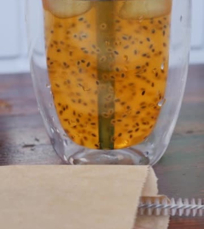 在一张木制桌子上放着一个玻璃杯，里面装有装有百香果种子的芒果饮料。玻璃中插入了可重复使用的蓝色吸管，