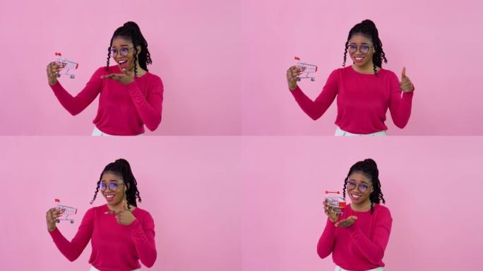 穿着粉色衣服的开朗年轻非洲裔美国女孩拿着玩具迷你购物车。少女家庭主妇初学者站在坚实的粉红色背景上