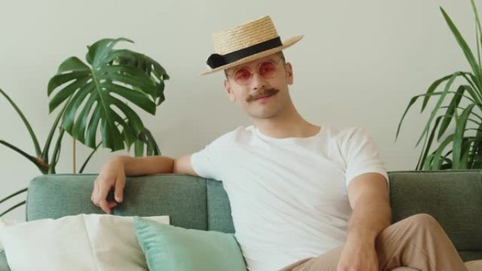 现代创意时尚潮人的肖像戴着小胡子，戴着帽子，圆形粉色太阳镜坐在沙发上，在现代阁楼公寓里眨眨眼。千禧一