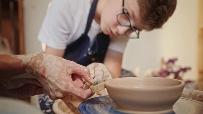 一个年轻的少年男孩坐在陶工的轮子上，在老师的帮助下，用特殊的工具在陶土上画了一幅画