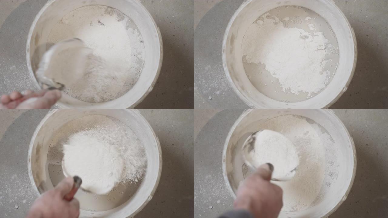 特写镜头，工匠的手将干燥的灰泥倒入一个装有水的容器中，以准备石膏混合物，以完成建筑物的维修和重建。房