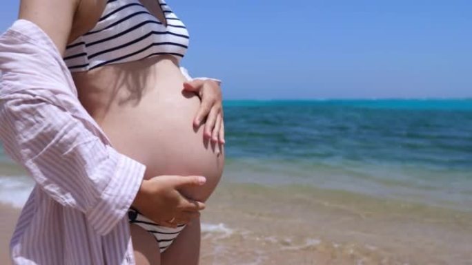 夏天快乐的年轻孕妇在海边，在天堂放松宁静的生育。一名妇女沿着海滩散步，在里面抚摸着婴儿的腹部