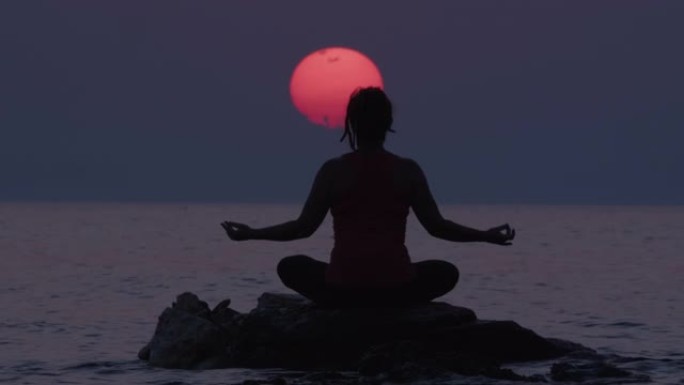 在令人惊叹的夕阳天空中，坐在海水中的石头上冥想的女人的剪影。治愈人的身体、精神和精神状态。