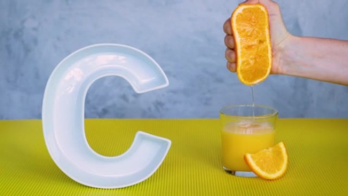 食品概念中的维生素c或抗坏血酸。人的手将橙子中的新鲜果汁挤压到灰色和黄色背景上的大陶瓷字母C附近的玻