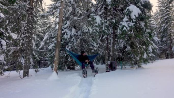 徒步旅行者带着雪鞋喝咖啡，躺在冬季森林的吊床上