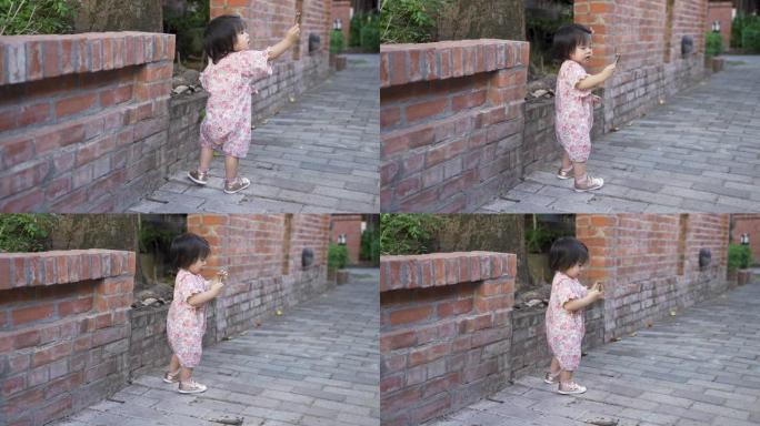 一个穿着日本和服的可爱的蹒跚学步的孩子的全长照片正在向远处的人展示手中的树皮，同时在红砖墙的文化公园