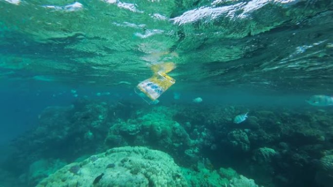慢动作，废弃的塑料包装在珊瑚礁附近的水面下慢慢漂流，热带鱼四处游动。黄色塑料袋漂浮在红海中
