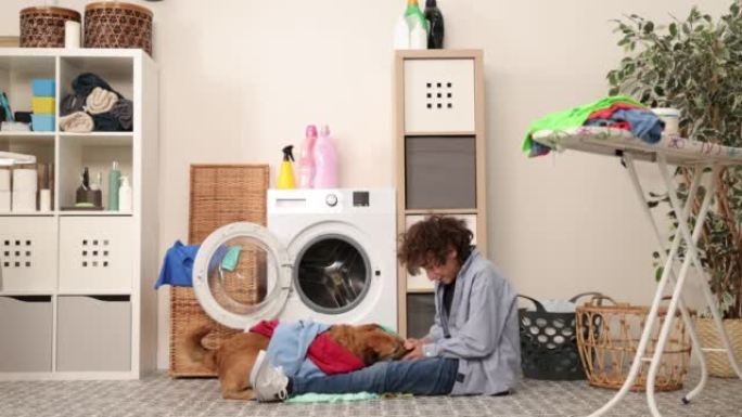 年轻的男孩穿着牛仔裤坐在洗衣机前。他用脏衣服给洗衣机装东西。洗衣房里的家务
