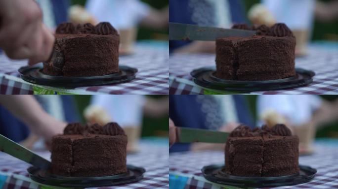 用刀在慢动作中近距离切片巧克力蛋糕。无法辨认的孩子在后院户外野餐庆祝生日。派对和童年的概念。