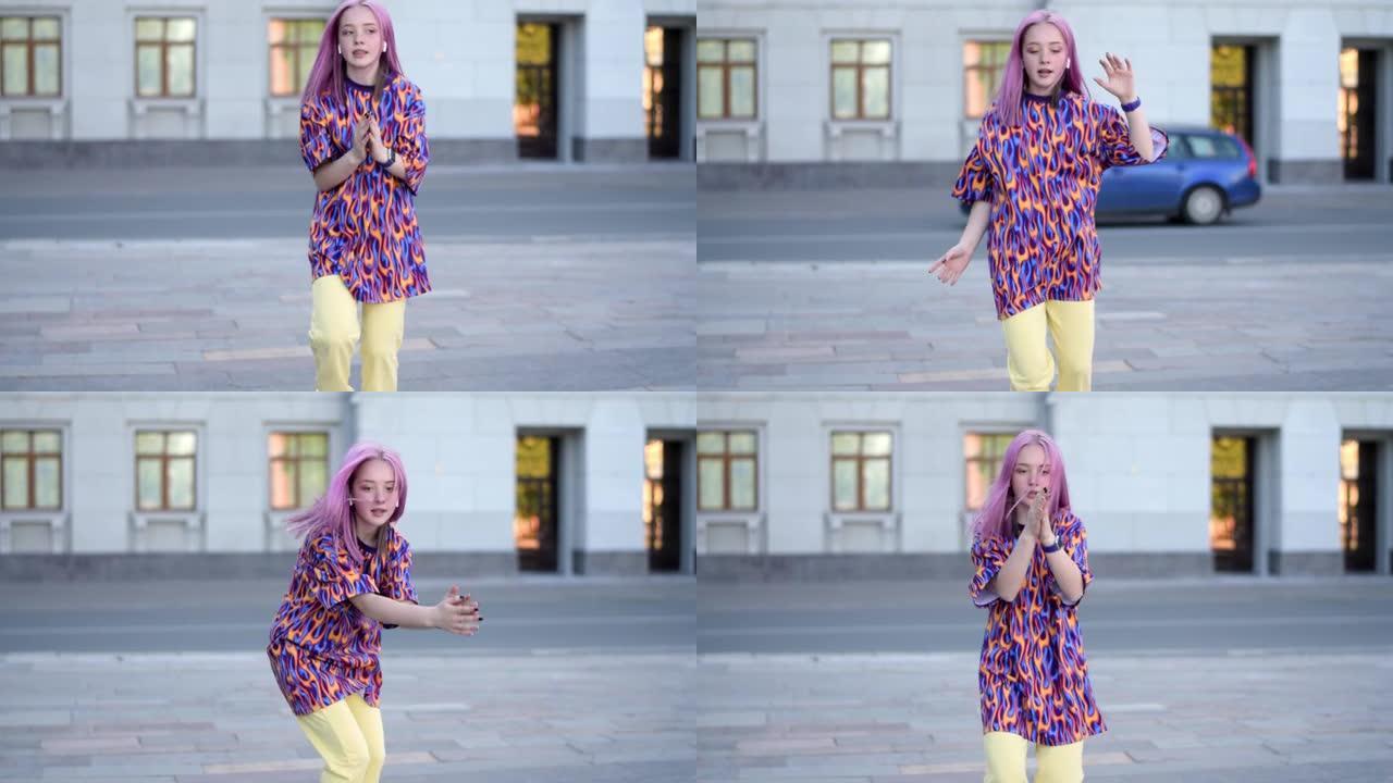 穿着五颜六色的明亮t恤和耳机的粉红色头发的十几岁时髦女孩在夏日的城市街道上跳舞嘻哈。Z世代。