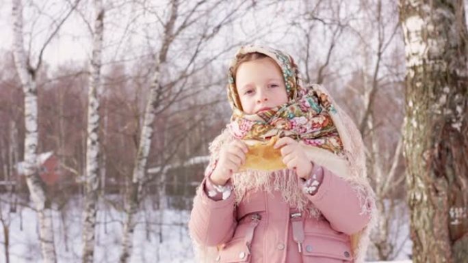 可爱的女孩在传统的俄罗斯头巾与百吉饼吃煎饼在冬天的背景。