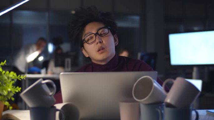 疲惫的商人在办公室晚上用电脑和咖啡杯在餐桌上睡着了