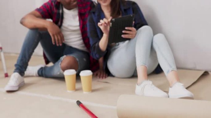 装修期间原始状态的公寓景观绘画墙壁油漆滚筒两个相爱的人坐在地板上喝咖啡，在平板电脑室浏览灵感，家具