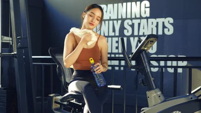 一名亚洲妇女在健身房休息，在健身在线小时用减肥锻炼机锻炼后，用毛巾擦干脸，喝矿泉水