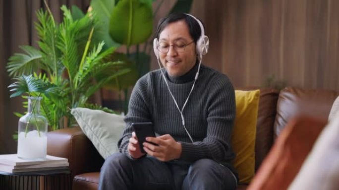 蛤蜊和安静的在家开朗的亚洲年轻男性带着耳机和智能手机播放列表选择在家里的客厅在智能手机上播放歌曲