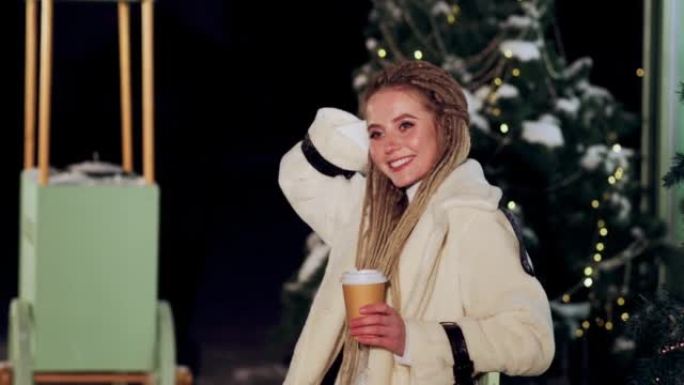 年轻漂亮的女人在圣诞树附近享受冬天晚上的热饮。茶杯或咖啡在手。