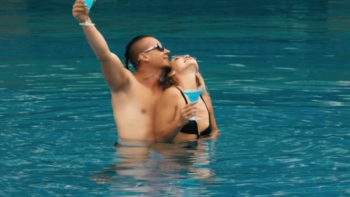 这对相爱的夫妇拥抱和亲吻，在室外酒店的游泳池里喝蓝色鸡尾酒酒精酒。高加索男人和女人的肖像