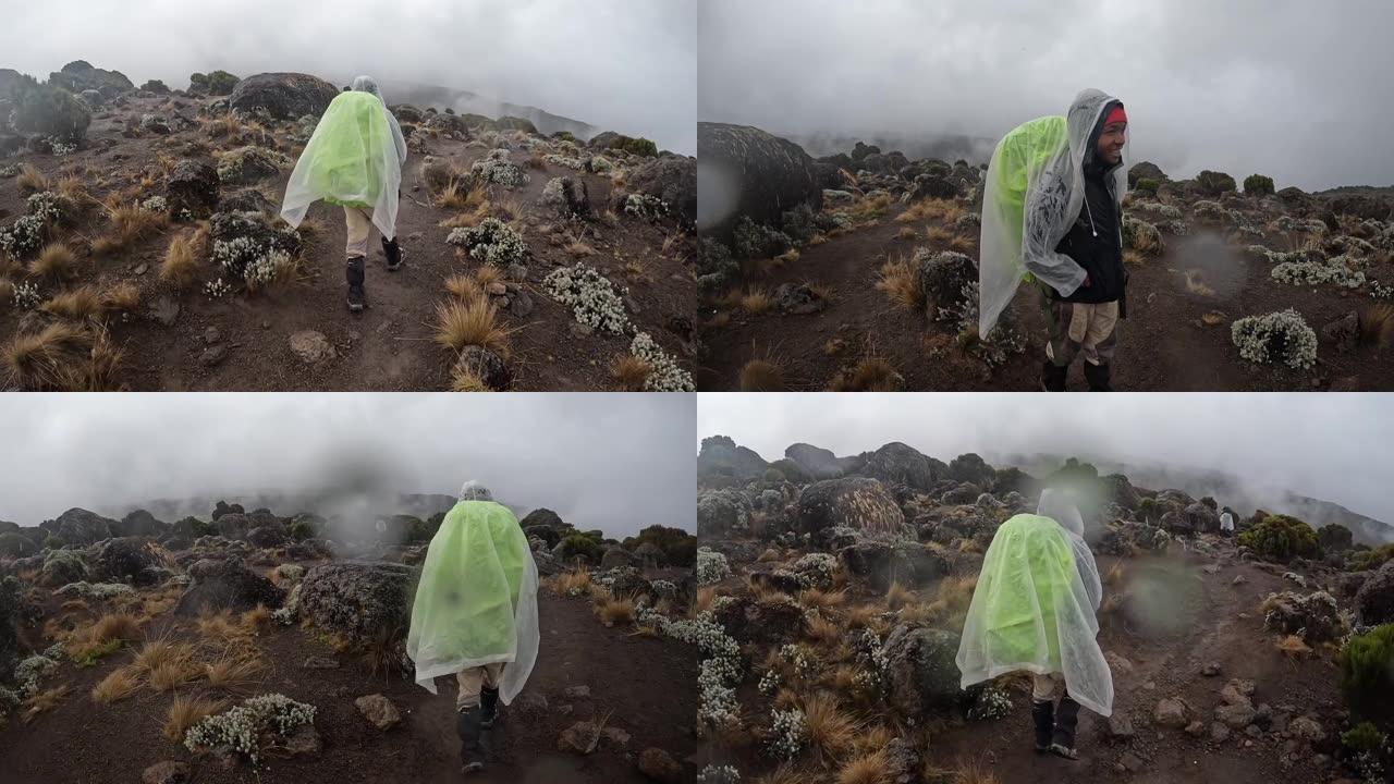 一个黑人背着大背包，穿着雨衣，沿着山路走着，经过了石头、巨石和灌木丛。山上有雨和雾。