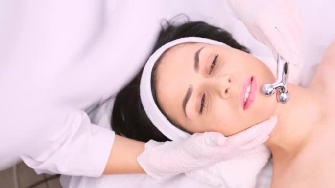 在美容诊所，护肤概念中，美容师在手套中使用化妆品Y形滚轮对女性进行面部提升按摩的裁剪照片。