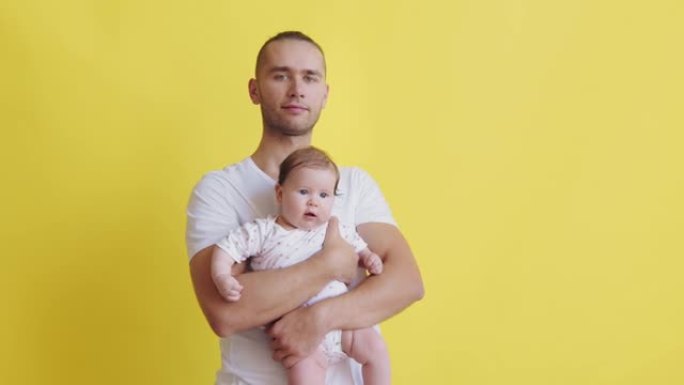 一位年轻英俊的父亲的肖像，怀里抱着他的小女儿，正看着孤立在黄色背景上的相机。千禧一代年轻家庭
