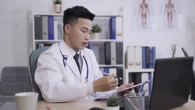 亚洲医务人员正在与患者的文件和手势交谈，同时在医院的办公桌上的笔记本电脑上进行虚拟医生的访问，远程医