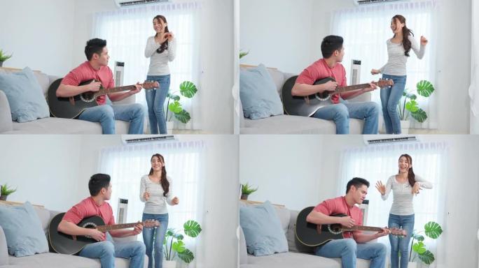 亚洲年轻的情侣在家里的客厅一起弹吉他。迷人的浪漫新婚，活跃的浪漫男人坐在沙发上，与美丽的女友跳舞演奏