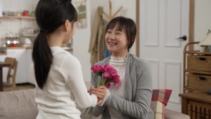亚洲女孩在家给妈妈送母亲节花的背景图。微笑的女人轻轻地抚摸着女儿的脸，一边闻到花束，一边说谢谢