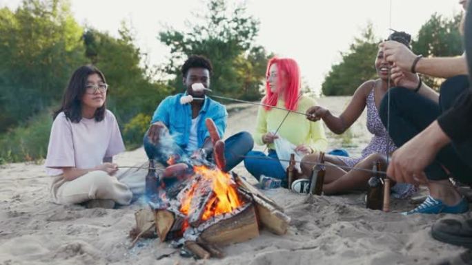 不同国籍的朋友坐在沙滩上，坐在沙滩上，喝啤酒，用火烧香肠，吃棉花糖，谈论在一起的时间，一起笑