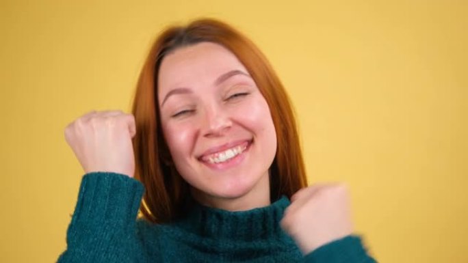 微笑着喜出望外的美丽姜黄色头发的年轻女子摆姿势做着赢家的手势，在工作室拍摄的黄色背景上孤立地说是。人