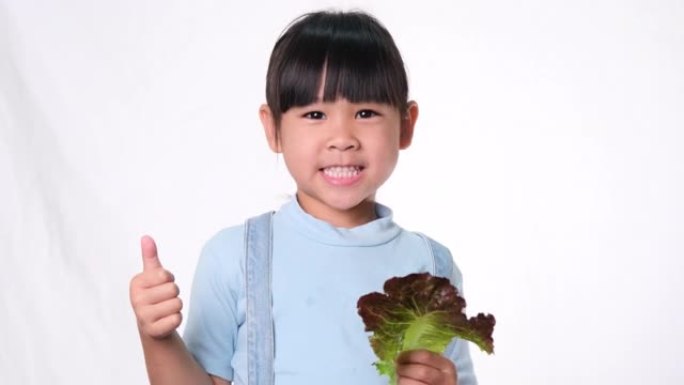 快乐的小女孩，带着新鲜的沙拉，在工作室的白色背景上竖起大拇指。儿童良好的健康习惯。医疗保健概念