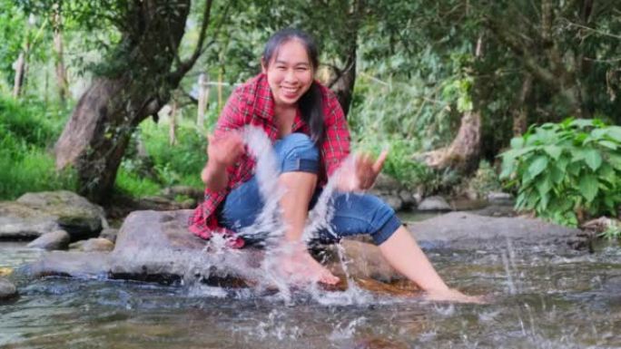 一位美丽的亚洲妇女坐在山林中的小溪中间的石头上玩水。假期和夏季概念。