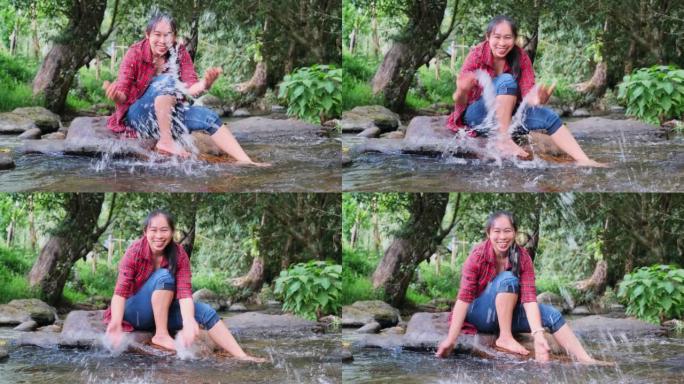 一位美丽的亚洲妇女坐在山林中的小溪中间的石头上玩水。假期和夏季概念。