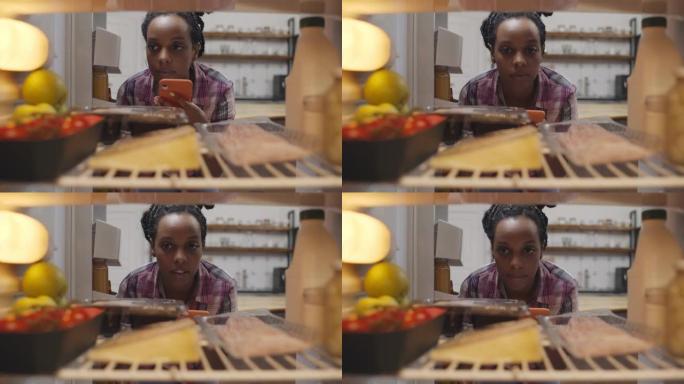 非洲裔美国女人在冰箱里看，在手机应用程序上做杂货店购物清单