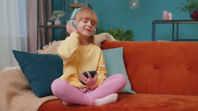 戴着耳机的智能手机的儿童女孩孩子在沙发上独自在家听音乐时跳舞