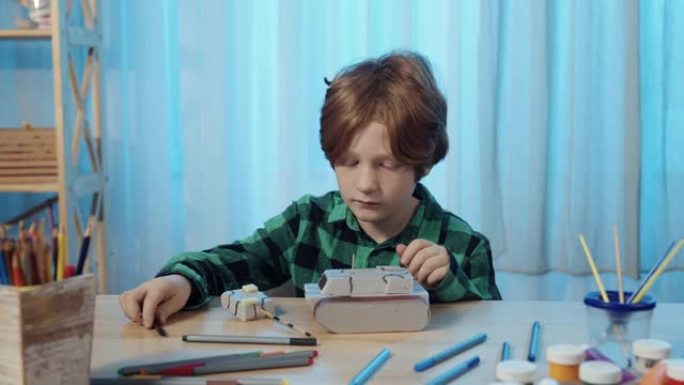 小男孩坐在房间的桌子旁，画玩具坦克。一个十几岁的男孩从事创造力，画画，爱好，发展，休闲。特写。慢动作