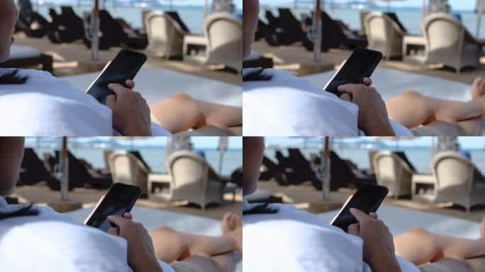 女人使用手机，赤脚交叉腿躺在海边的沙滩床椅子上