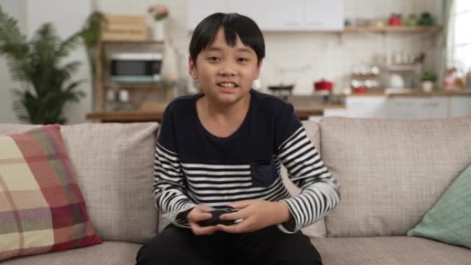 亚洲男孩在家玩电子游戏的慢动作，输了。悲伤的男生坐在沙发上，手里拿着操纵杆在家里的客厅。