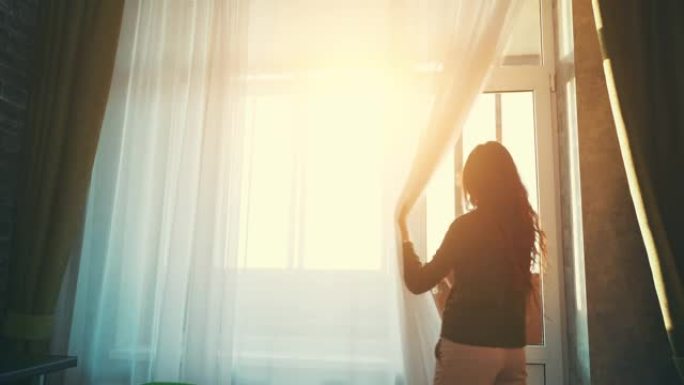 长发的年轻女孩或女人的后视接近大窗户，在夕阳的阳光下打开窗帘