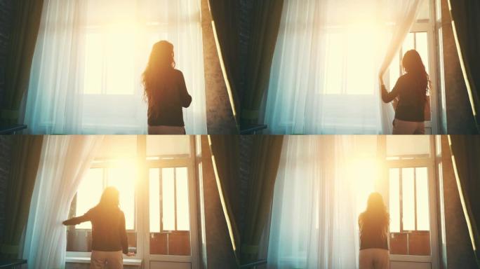 长发的年轻女孩或女人的后视接近大窗户，在夕阳的阳光下打开窗帘
