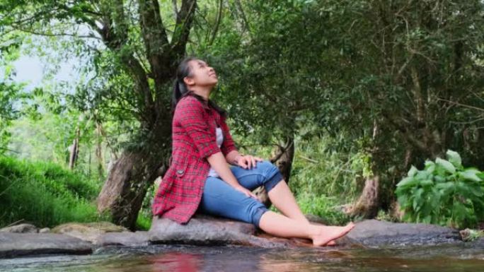 一位美丽的亚洲妇女坐在山林中的小溪中间的石头上，享受大自然。假期和夏季概念。