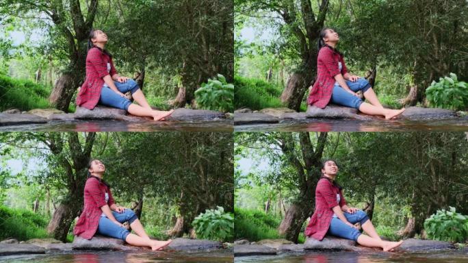一位美丽的亚洲妇女坐在山林中的小溪中间的石头上，享受大自然。假期和夏季概念。