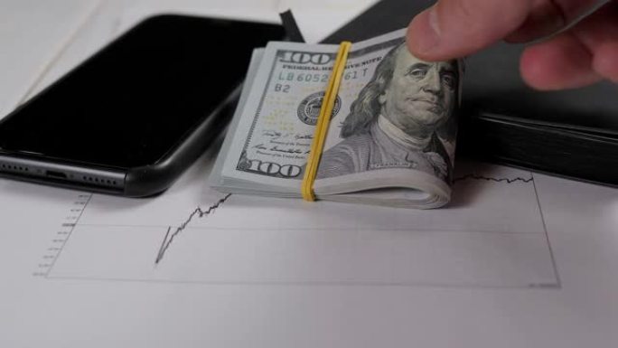 一个男人把一叠钞票放在桌子上，桌子上放着电话和财务图表。