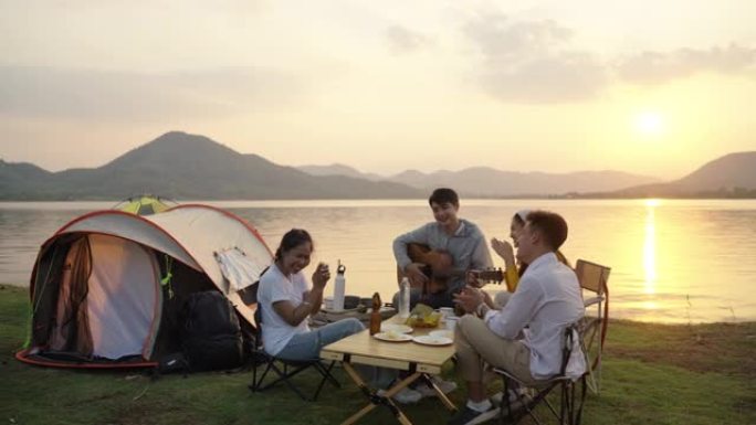 亚洲四个年轻朋友在河边露营野餐，他们聊天，大笑，喝酒庆祝啤酒。休闲装，夏天的时候，夕阳的亮光。