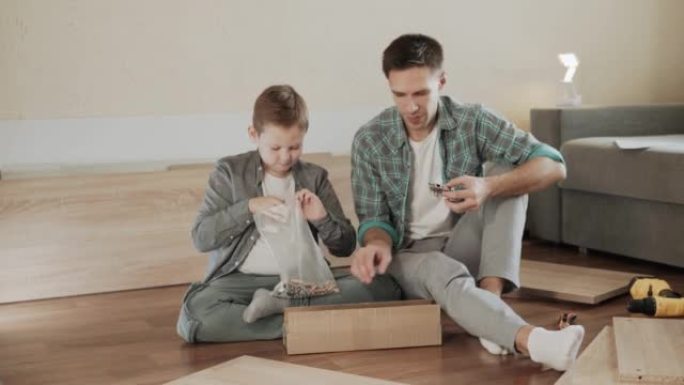 爸爸和儿子坐在地板上，拿出配件从盒子里组装橱柜。