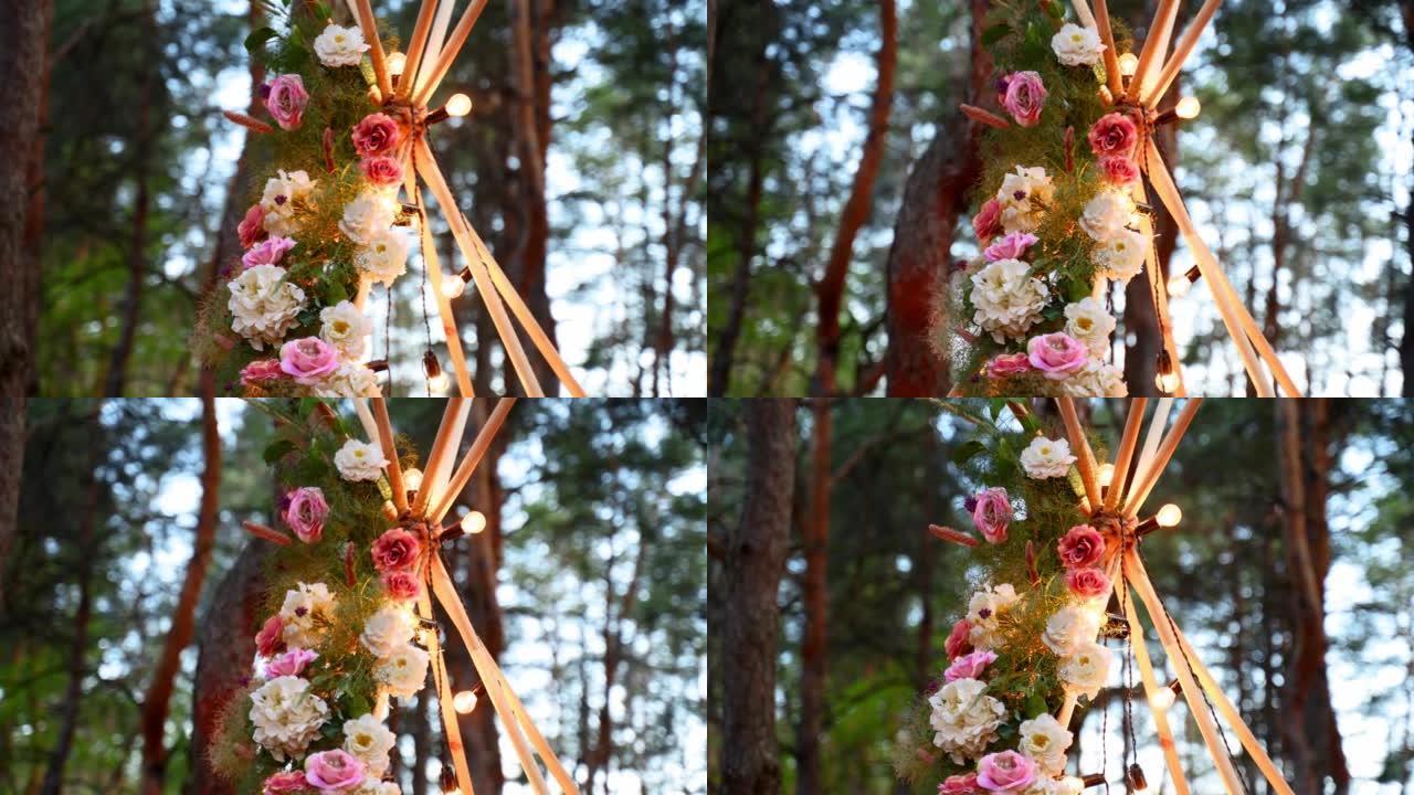 波希米亚的提皮木制拱门装饰有粉红玫瑰，地毯上的蜡烛，潘帕斯草，晚上在松林的户外婚礼仪式场地包裹着仙女