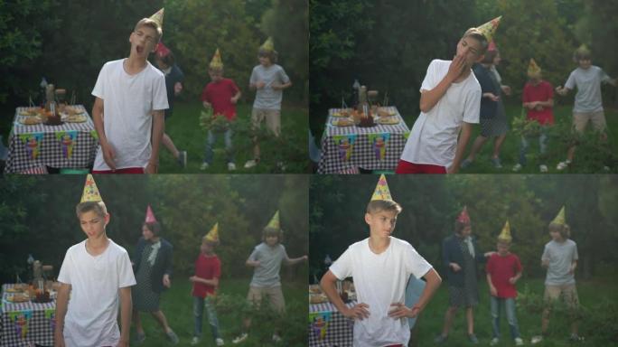 无聊的十几岁男孩在生日聚会上打哈欠的肖像，模糊的一群朋友在背景中跳舞。毫无兴趣的高加索少年在春夏公园