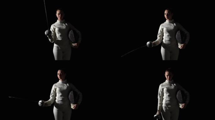 一名年轻女子击剑手的肖像用剑杆致敬。穿着白色制服的运动员在黑色背景的黑暗工作室中摆姿势，并带有舞台灯