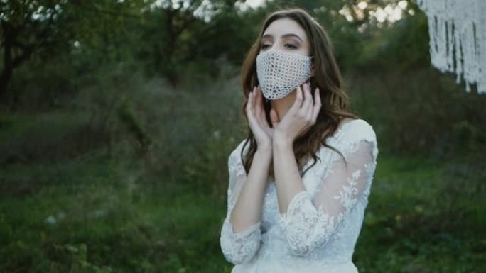戴着面具摆姿势的新娘肖像。公园里穿着婚纱的漂亮年轻新娘。她的脸上有一个带鹅卵石的时尚白色面具
