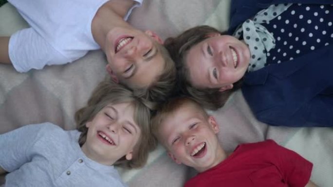 在春夏公园，轻松快乐的孩子笑着看着躺在毯子上的相机的高角度视角肖像。无忧无虑的高加索男孩和女孩在户外